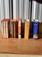 Several new books standing upright on a wooden surface with a metal background, including a three volume set titled 'The Great Writers', 'The Dead Sea Scrolls', 'Pagan, Royal, and Christian', and 'The Oxford Dictionary of Quotations'.