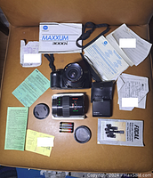 Minolta Maxxum 3000i camera with 28-70mm lens attached, 70-210mm lens beside, original box and manuals, camera case, lens caps, batteries, and other documentation on brown surface.