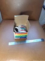 Mug inside original cardboard box, showing packaging and size reference with ruler.