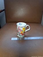 Ceramic mug with multicolor floral print and botanical flower names on front placed on brown chair with ruler for size reference.