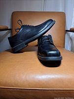Side and angled view of two black leather lace-up dress shoes on tan leather chair, showing upper shape and design.