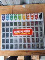 Top view of the closed box showing 50 metal puzzles in a plastic insert with colorful category labels on the box lid