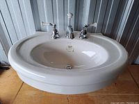 Oval white ceramic American Standard washbasin with chrome triple-handle faucet on wooden floor.