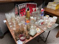 Table with assorted glassware including clear and tinted glasses, decanters, mugs, ashtrays with dice and playing cards visible in background