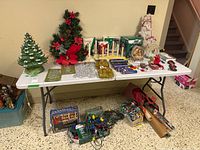 Table showing entire lot of Christmas decorations including ceramic trees, candles, lights, snow globe, paper and plastic ornaments.