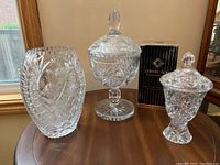 Full view of the three crystal pieces displayed on a wooden table: vase with floral etching, 10-inch lidded chalice, 15-inch lidded candy dish, and a box behind