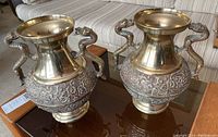 Pair of brass vases shown side by side on a glass table, highlighting overall shape and design including dragon handles and flared rims.
