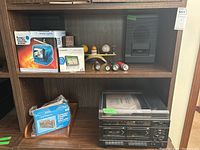 View of two shelves with various electronics including Soundesign stereo components, portable TV, Garmin GPS, cassette recorder, brass candleholder, mini flashlights, baseball and desk tray