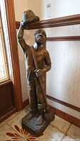 Front angle view of the full wooden statue showing the man holding a hat raised in his right hand and a broom in his left hand, barefoot, wearing a short-sleeve shirt and pants.