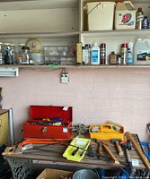 Photo of red toolbox open with assorted hand tools and yellow organizer on table, saws and hammers nearby, shelves above with cleaning products and hardware jars.