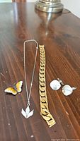 Jewelry lot displayed on wooden surface: gold tone chunky men's bracelet, silver necklace with flower pendant, butterfly brooch, and pair of round silver cufflinks.