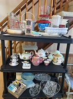 Overall view of three shelves with various teapots, measuring cups, condiment plates, and kitchenware arranged on them.
