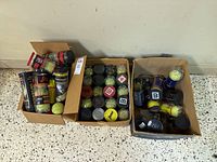 Photo showing three cardboard boxes each filled with multiple unopened cans of tennis balls of various brands and colors.
