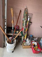 Garden tools including broom, rakes, umbrellas, and various handled tools standing in bucket and leaning against wall