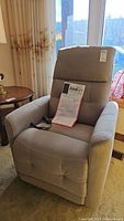 Front-left view of grey upholstered power lift recliner with instruction manual and remote on seat