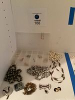 Photo of assorted jewelry laid out on white surface including beaded necklaces, brooches, clip earrings, and small compartments with jewelry pieces.