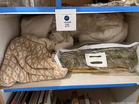 Four blankets on shelf including a beige and white patterned knitted blanket, white down comforter with pillow, an off-white blanket, and a green blanket in a clear plastic bag