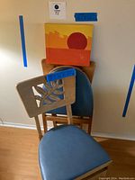 Two wooden card table chairs with blue vinyl seats and cut-out leaf design backs stacked in front of a wall with a small painted canvas of a red sun.