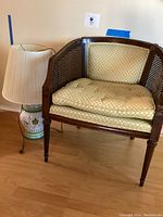 Wood framed chair with woven cane sides and cushioned seat and back next to floral ceramic lamp with cream pleated shade