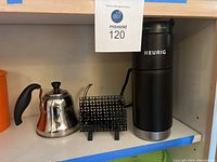Keurig coffee machine in black next to small silver teapot with black handle and long spout and black metal pod holder on white shelf