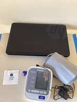 Closed HP black laptop with visible HP logo on lid, accompanied by an Omron blood pressure monitor with gray cuff and attached tubing, with a small paper tag numbered 226.
