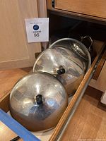 Two large stainless steel pot lids with black plastic knobs inside kitchen cabinet