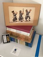 Photo shows the signed painting of three dancing religious Jewish men above stacks of Passover Haggadah books and a packaged car mezuzah