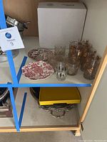 Photo showing the collection of glasses and two salad plates, including glassware with gold floral patterns and black/clear striped glass, on a shelf.