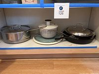 Photo of various kitchen items including a white salad spinner, cast iron skillet with lid, and stainless steel pan with lid placed on shelf