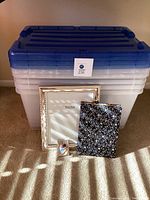 Five large clear plastic storage bins stacked with blue lids, a silver picture frame, small oval photo frame, and floral journal placed in front of the bins.