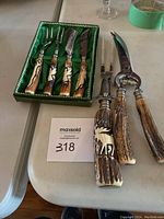 Overview of seven carving and serving utensils: four small forks and knives in green presentation tray, plus carving knife, carving fork, and poultry shears with antler-style handles