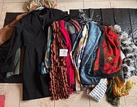 Scarves and black jacket displayed laid out on the floor on black mats, show multiple scarves with different colors, textures and patterns alongside a black hooded jacket.