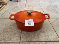 Orange cast iron pot with lid and two side handles shown from a front angle on tiled floor.