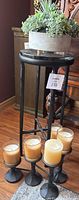 Photo showing the black metal plant stand with artificial succulent plant in grey round pot on top, next to black metal candle holder with glass-contained candles on floor.