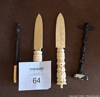Four carved items displayed on a brown surface including two ivory-colored letter openers, a black carved pen with gold tip, and a black carved totem pole statue on a base with chipped corner.