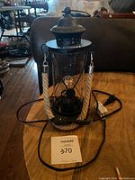 Full front view of the lantern lamp showing its black aluminum top and base, clear glass body, natural rope handle with metal caps, power cord, and LED filament bulb inside.