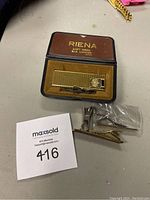 Gold-tone Riena piezo gas lighter in brown case on table with additional metal clips and pins nearby.