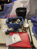 Photo showing the bulk of office supplies including notebooks, blue plastic bin with assorted items such as pencil sharpener, stapler, paper clips, tape, and a black octagonal garbage bin behind the bin