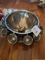 Large metal punch bowl on matching plate, surrounded by 12 metal cups hanging on hooks around bowl rim with ladle inside bowl.