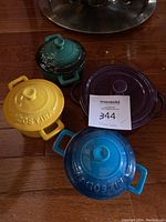 Four small stoneware casserole dishes with lids displayed on wood floor. Three are Lagostina brand in yellow, blue and green. One is purple VE stoneware.