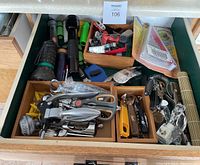Top-down photo of a drawer filled with assorted kitchen utensils and several flashlights. Visible are multiple corkscrews, can openers, scissors, a jar opener in original packaging, chip clips, corn holders, and various flashlights.