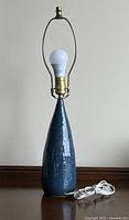 Full view of blue ceramic table lamp with harp, light bulb, and power cord on wooden surface.