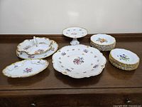 Image showing an assortment of porcelain plates and serving dishes with floral patterns and gold trim displayed on a wooden surface.