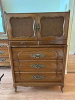 Front view of the wooden high boy dresser showing the upper cabinet doors with cane panels, two small drawers, and three large drawers with ornate metal handles.