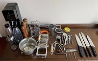Full view of the lot on a wooden surface showing the soda machine, pepper mills, metal graters, potato ricer, kitchen knives, and various small utensils arranged neatly.