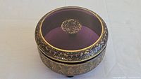 Top view of round purple glass box with gold oroplastic decoration on lid and finial knob.