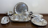 Full lot of silverplate serving pieces arranged on surface, showing large round platter, rectangular footed platter, tea pot, ice bucket, sugar and cream set, salt and pepper shakers, cover dish and more.