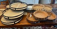 Photo showing 5 farmhouse round wooden serving trays with black metal frames stacked and 2 rattan scallop trays, one larger and one smaller, with natural wooden bases and woven rattan edges placed on table.