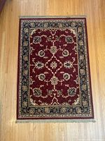 Top view of rectangular area rug showing red floral patterned field with navy and cream border on wooden floor.