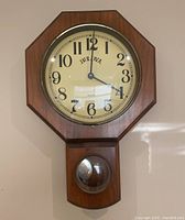 Front view of Bulova wall clock showing octagonal wood frame, cream dial with black numerals, and pendulum behind glass in lower compartment.